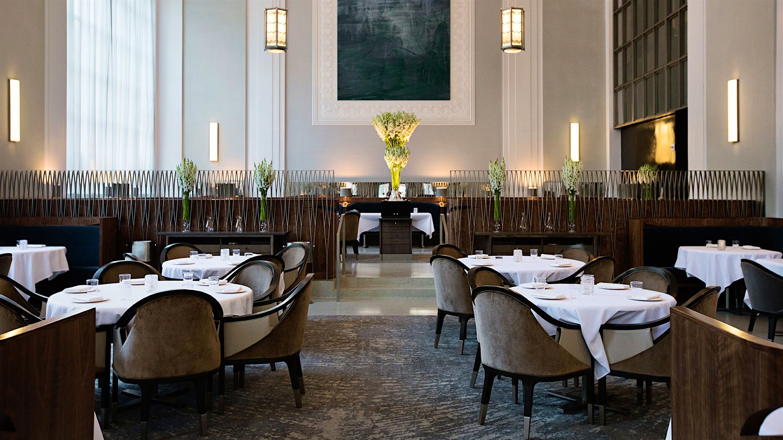 The dining room at Eleven Madison Park, with a large gray painting, glass vases of flowers and wood-cloth chairs